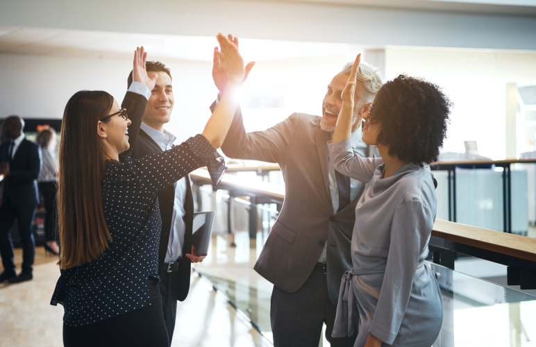 Diverse group of smiling businesspeople high fiving each other while standing together in a corridor of a modern office building